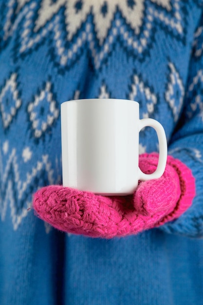 Tazza bianca di Natale finta in mano su una donna che indossa guanti rosa e maglione blu.