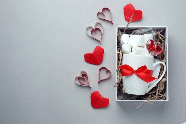 Tazza bianca, dolcezza e decorazioni rosse in confezione regalo su sfondo grigio