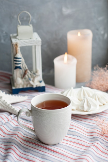 tazza calda di tè nero con un piatto di ariose meringhe su una tovaglia a strisce, stelle marine decorative e lanterna, candele di cera, erbe decorative