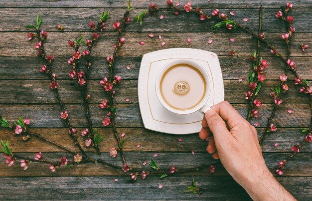 Tazza con il latte del caffè in una mano maschio sulla a di una vecchia tavola di legno con i rami della pesca con i fiori rosa, vista superiore