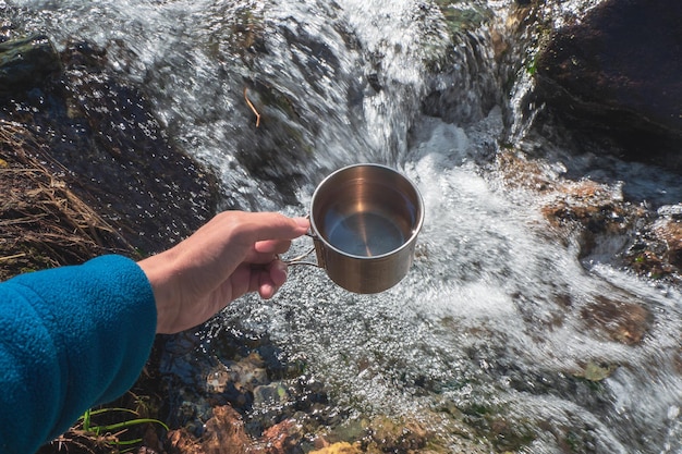 Tazza con pura acqua glaciale di un ruscello di montagna. Mano che tiene una tazza di escursionisti con acqua potabile pulita.