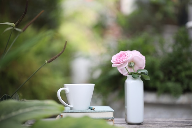 Tazza da caffè bianca con rosa rosa in vaso di ceramica e quaderni su tavolo di legno stagionato all'aperto