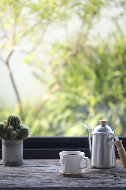 Tazza da caffè bianca e caffettiera in acciaio inossidabile con cactus sul tavolo davanti alla finestra