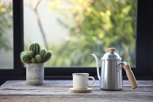 Tazza da caffè bianca e caffettiera in acciaio inossidabile con cactus sul tavolo davanti alla finestra