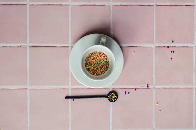 Tazza da caffè bianca piena di granelli di caramelle e un cucchiaio nero su uno sfondo di piastrelle rosa