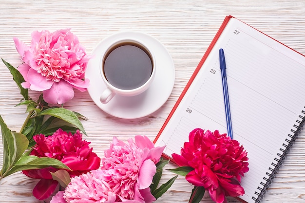 Tazza da caffè del mattino per colazione, taccuino vuoto, matita e fiori di peonia rosa su pietra bianca vista da tavolo in stile piatto laico.