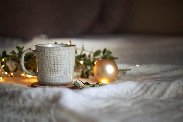 Tazza da caffè in ceramica su fondo a maglia beige con foglie di eucalipto e luci di ghirlande natalizie, abbraccio a casa