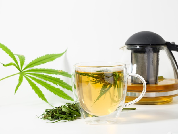 Tazza da tè alla cannabis con foglie di marijuana e fiori in tazza di vetro, bollitore marrone olio CBD su bianco