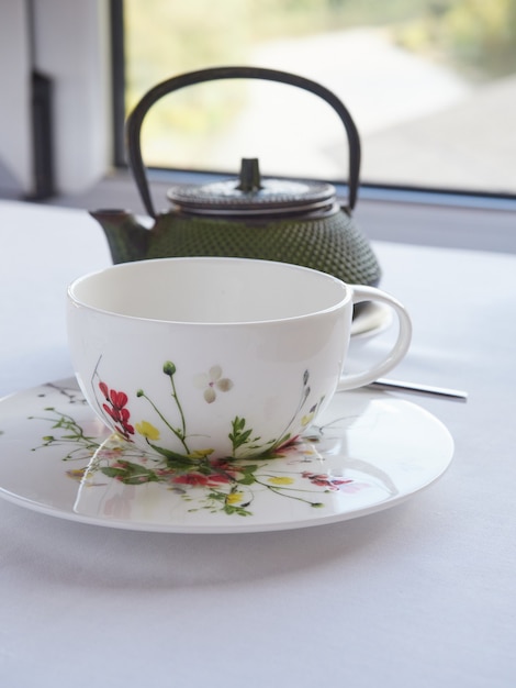 Tazza da tè bianca decorata con fiori dipinti e teiera orientale davanti a una finestra bianca