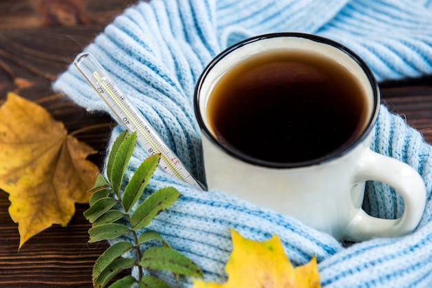 Tazza da tè con termometro, sciarpa blu e foglie di autunno su fondo in legno. Stagione influenzale in autunno, malattia.