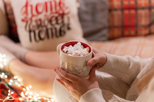 Tazza di cacao o bevanda al caffè con marshmallow in donna che si tiene per mano di donna in maglione lavorato a maglia rilassarsi e godersi le vacanze in un letto comodo e accogliente a casa alla vigilia di natale o capodanno