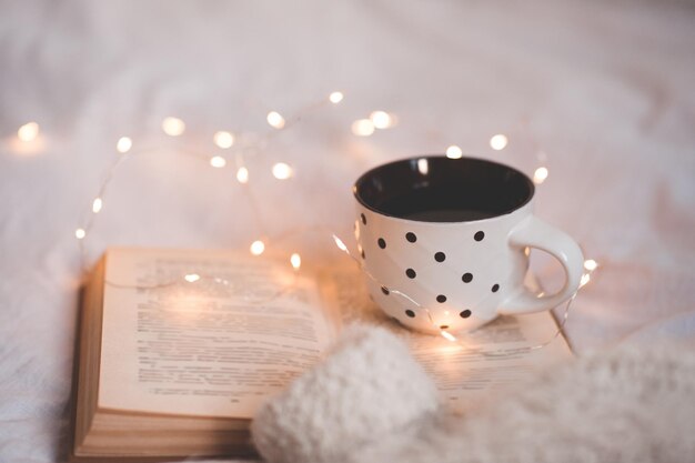 Tazza di caffè caldo che resta sul libro di carta aperto sopra il primo piano delle luci d'ardore. Ora di colazione. Buon giorno. Stagione invernale.