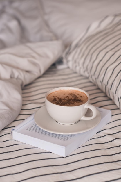 Tazza di caffè caldo con cioccolato in cima che resta sul libro di carta nel primo piano del letto. Buon giorno. Colazione.