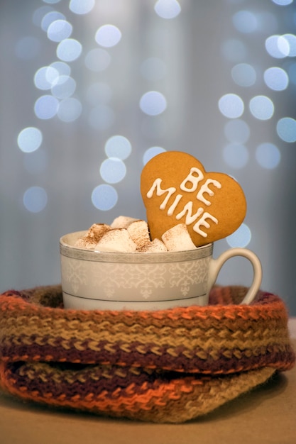 Tazza di caffè caldo e aromatico con marshmallow e biscotto al cuore con frase Be Mine su lucine blu