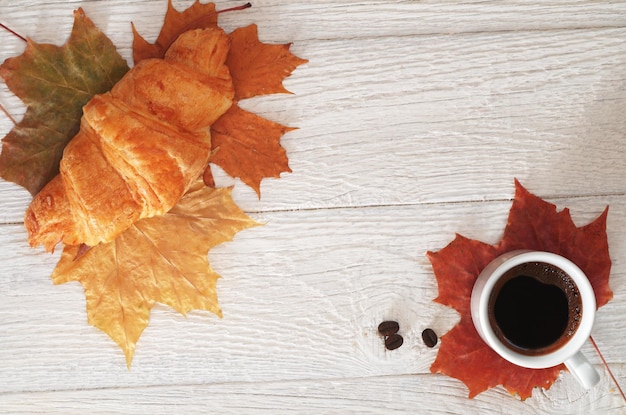 Tazza di caffè caldo e croissant sulla tavola di legno bianca con la vista superiore delle foglie di autunno