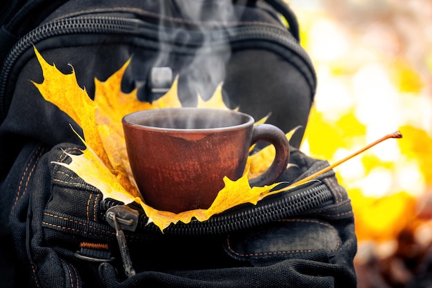 Tazza di caffè caldo e foglia d'acero su uno zaino nella foresta autunnale