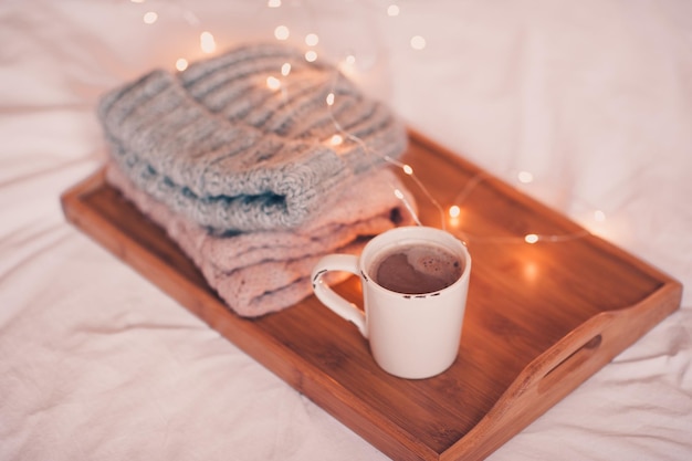 Tazza di caffè che rimane sul vassoio di legno con cappello lavorato a maglia e sciarpa da vicino sopra le luci di Natale Buongiorno Stagione invernale