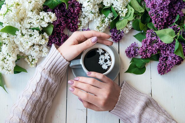 Tazza di caffè con fiori lilla e mani femminili piatte con sfondo bianco caffetteria