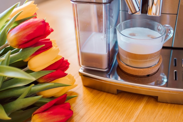 Tazza di caffè con latte e fiori di tulipani sul tavolo da cucina in legno Espresso cappuccino appena preparato nella macchina del caffè per la colazione Arredamento interno elegante per la casa