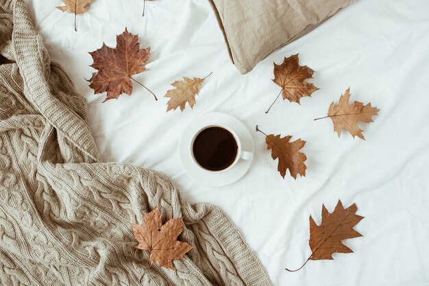 Tazza di caffè con latte, foglie secche sul letto con lenzuola bianche, cuscino, coperta