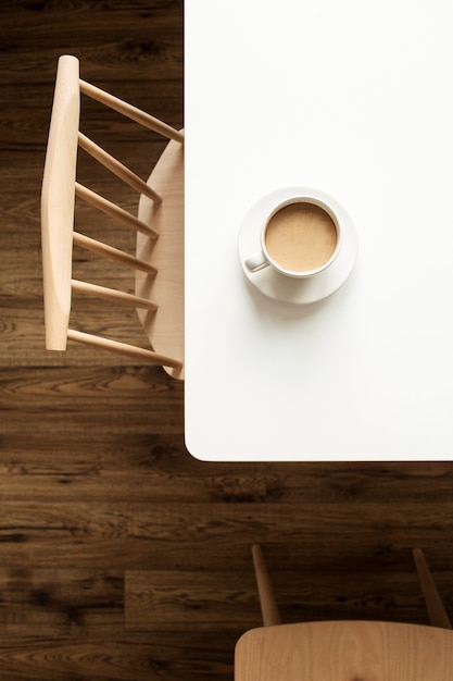 Tazza di caffè con latte sul tavolo bianco vicino alla sedia di design e pavimento in legno