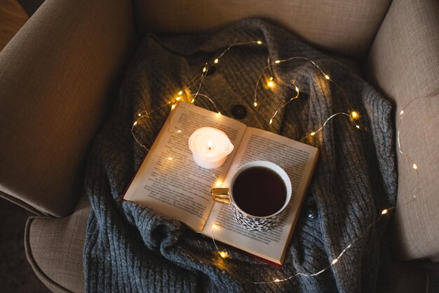 Tazza di caffè con libro aperto di carta e candela profumata nella sedia domestica sopra la vista superiore delle luci di Natale