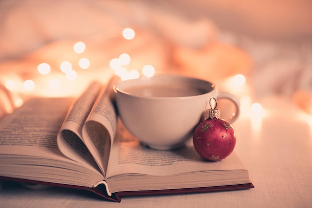 Tazza di caffè con libro aperto e pagine a forma di cuore sopra le luci di Natale da vicino. Vacanze invernali.