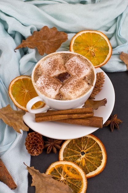Tazza di caffè con marshmallow, cacao, sciarpa, foglie, arance secche, spezie, su sfondo grigio. Deliziosa bevanda autunnale calda, umore mattutino. copyspace.