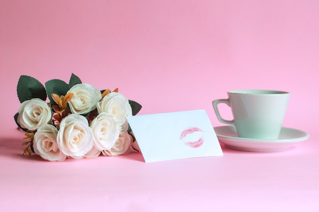 Tazza di caffè con rose e bacio sulla busta bianca isolata su sfondo rosa