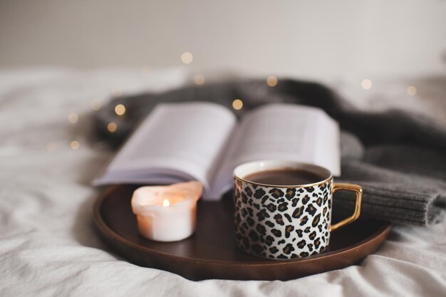 Tazza di caffè con stampa leopardata che rimane su vassoio di legno con candela accesa profumata e libro di carta aperto su tessuto lavorato a maglia a letto a casa Buongiorno Orario di colazione Accogliente atmosfera hygge