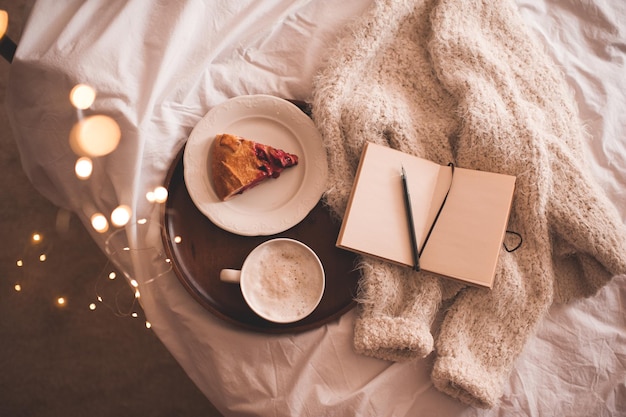 Tazza di caffè con torta di frutta, tessuto a maglia e taccuino a letto con luci di Natale da vicino. Vista dall'alto. Stagione delle vacanze invernali.