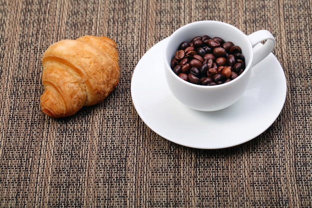 Tazza di caffè con un cornetto e chicchi di caffè freschi su uno sfondo marrone