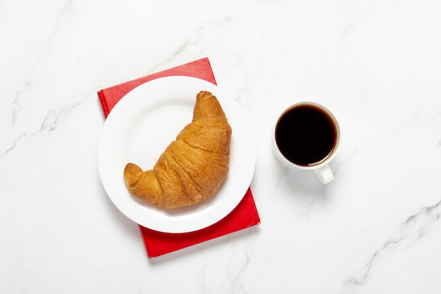 Tazza di caffè, croissant su un piatto e un libro su un tavolo di marmo