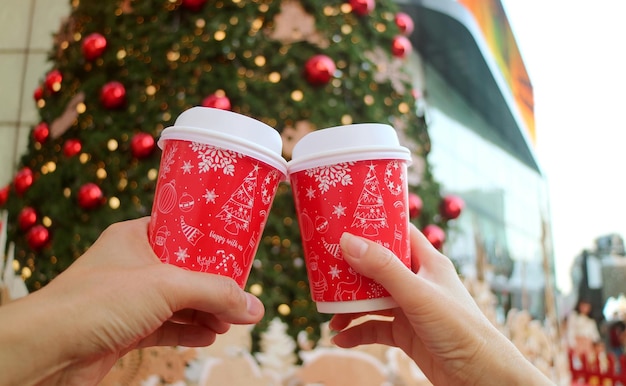 Tazza di caffè da asporto a tema natalizio nelle mani di una coppia contro l'albero di Natale sfocato