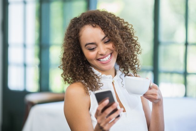 Tazza di caffè della bella tenuta della donna e telefono cellulare usando