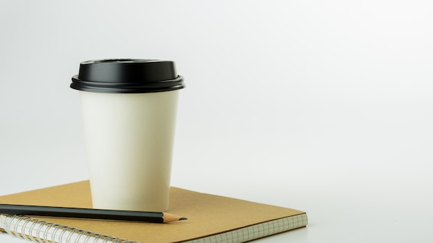 Tazza di caffè di carta e un taccuino sul fondo bianco dello scrittorio con lo spazio della copia. - forniture per ufficio o concetto di educazione.