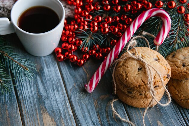 Tazza di caffè e biscotto con gocce di cioccolato mescolati con decorazioni festive per le vacanze invernali. spirito natalizio e concetto di svago. stringa di perline rosse su ramo di abete e bastoncino di zucchero.