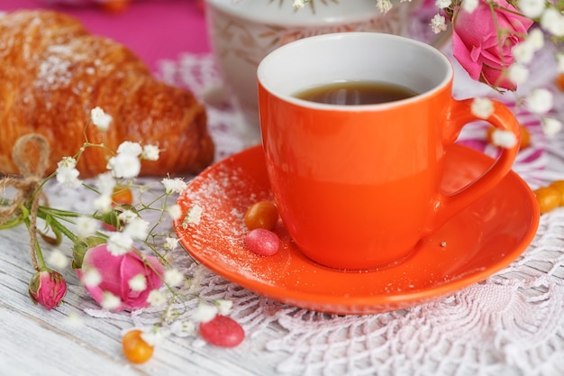Tazza di caffè e croissant sono decorati da tovaglioli, rose e caramelle su un tavolo di legno bianco.