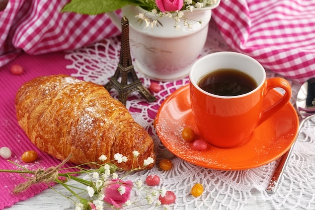 Tazza di caffè e croissant sono decorati dalla piccola Torre Eiffel, tovaglioli, rose e caramelle su un tavolo di legno bianco