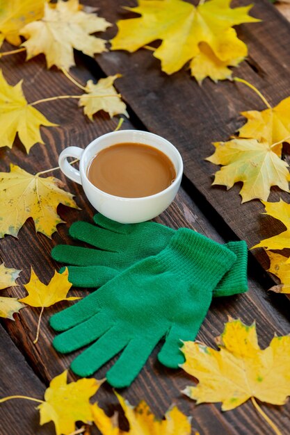 Tazza di caffè e guanti verdi accanto alle foglie di acero sulla tavola di legno.