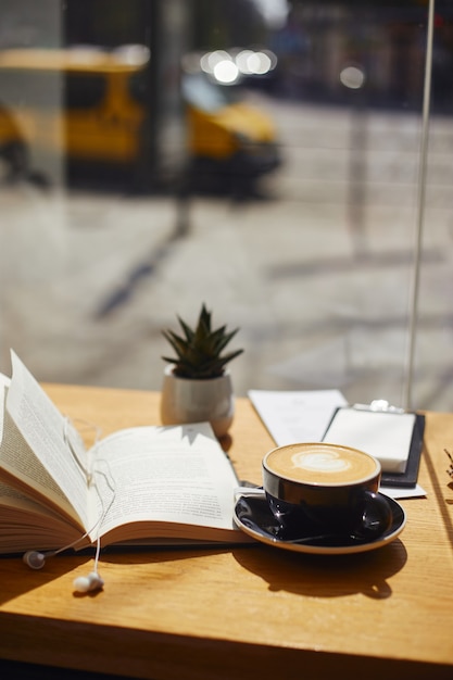 Tazza di caffè e libro sul tavolo nella caffetteria vicino alla finestra