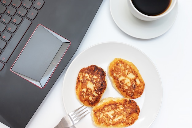 tazza di caffè e un piatto con cagliata, frittelle di ricotta per la colazione sul tavolo vicino al computer portatile