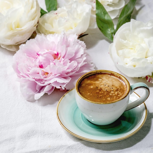 Tazza di caffè espresso, fiori di peonie rosa e bianchi con foglie su sfondo bianco in tessuto di cotone. Disposizione piatta, copia spazio. Immagine quadrata