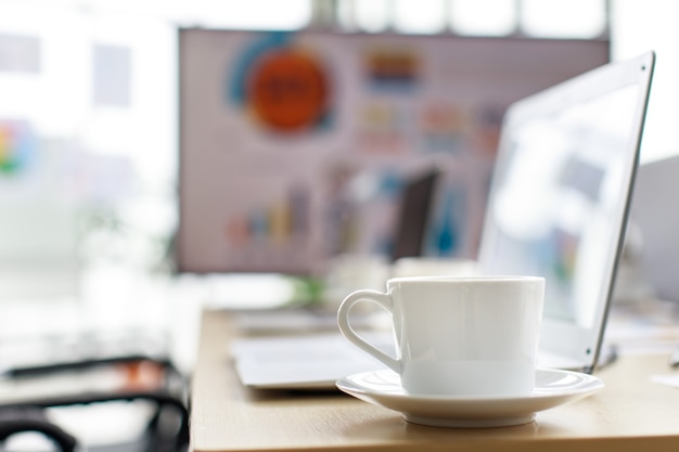 Tazza di caffè gustosa bianca calda su un piccolo piatto posizionato sul tavolo di lavoro con computer portatile nella sala riunioni davanti al grafico del tasso di crescita target e al rapporto grafico sul monitor del grande schermo in sfondo sfocato.