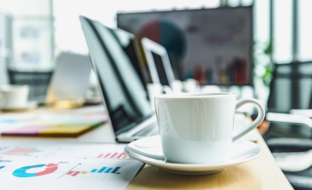 Tazza di caffè gustosa bianca calda su un piccolo piatto posizionato sul tavolo di lavoro con computer portatile nella sala riunioni davanti al grafico del tasso di crescita target e al rapporto grafico sul monitor del grande schermo in sfondo sfocato.