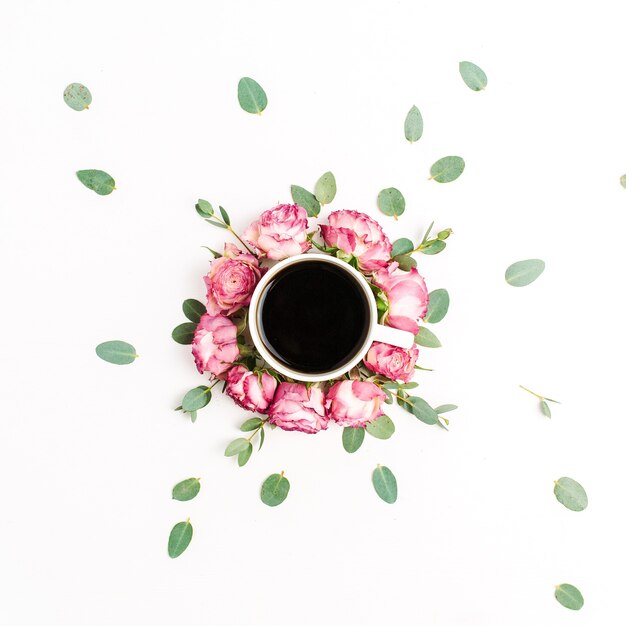 Tazza di caffè in cornice di boccioli di fiori rosa rosa e rami di eucalipto su sfondo bianco. Disposizione piatta, vista dall'alto