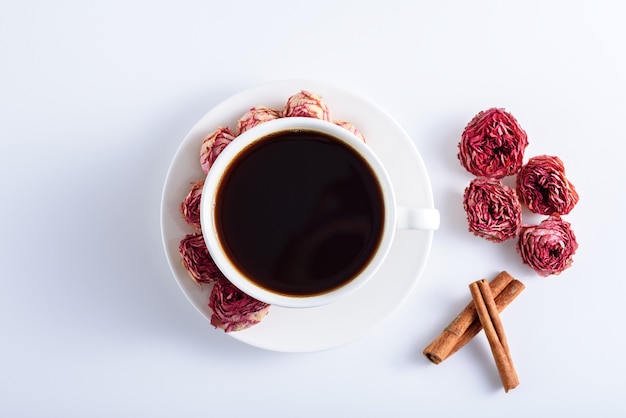 Tazza di caffè nero con rose sulla piastra, bastoncini di cannella sul tavolo bianco