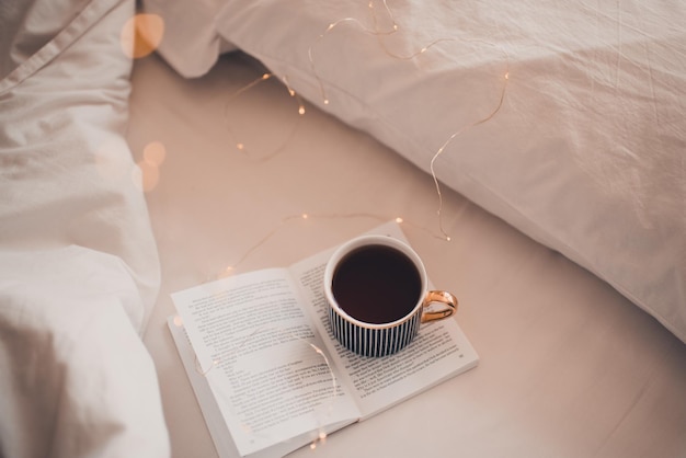 Tazza di caffè nero rimanere su un libro di carta aperto a letto su una coperta bianca primo piano Accogliente mattina