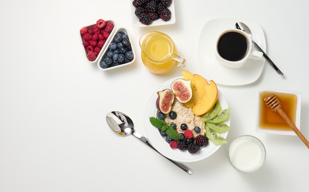 Tazza di caffè nero, un piatto di farina d'avena e frutta, miele e un bicchiere di latte su un tavolo bianco, una sana colazione mattutina, vista dall'alto