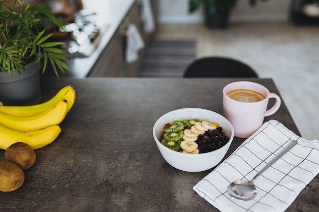 Tazza di caffè rosa, ciotola con kiwi e banana tritati di frutti tropicali, mirtilli, cucchiaio sul bancone del bar in elegante cucina loft.